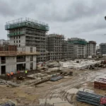 Chantier de construction avec plusieurs immeubles encours, grues et ouvriers en sécurité sur le terrain humide, piles de matériaux et zones de travail visibles. pénurie main d'oeuvre btp.