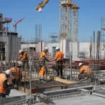 Chantier de construction avec ouvriers en vêtements orange et casques, ferraillage et grues en arrière-plan, illustrant les métiers du btp qui recrutent.